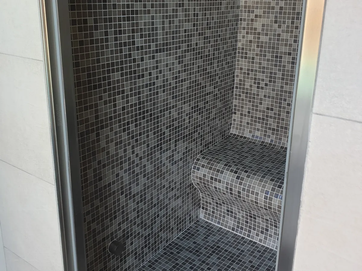 Hammam en mosaïque grise vu par sa porte vitrée à cadre inox : banquette, grille d'évacuation au sol