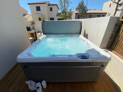 Spa installé sur une terrasse en bois entre deux murs, couverture relevée, maisons à l'arrière-plan