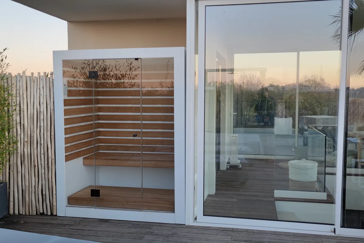 Sauna vitré à cadre blanc installé sur une terrasse en bois, au coucher du soleil